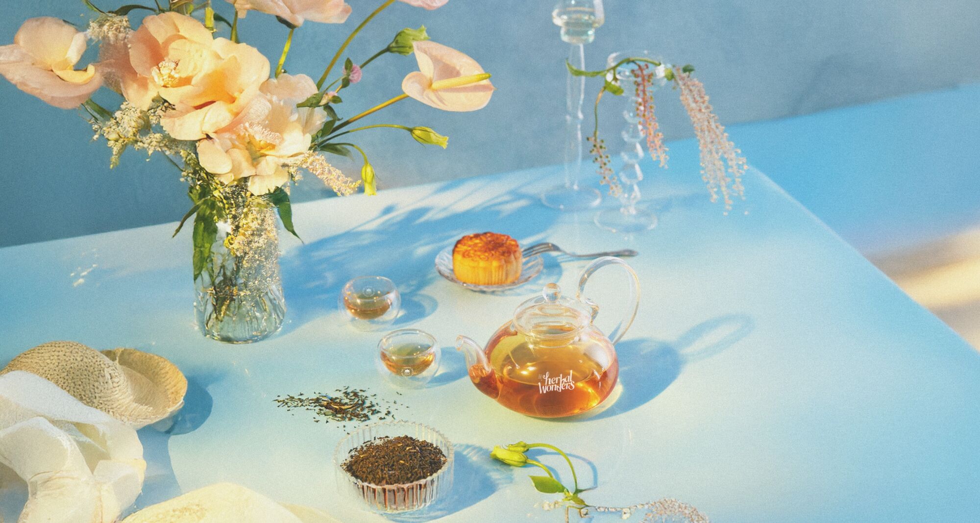 A glass teapot of brewed tea, cups, loose leaves and a cake on a sunlit blue table